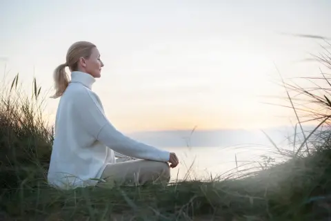 Kobieta w białym swetrze medytuje na plaży, szukając spokoju i sposobu, jak oczyścić umysł.