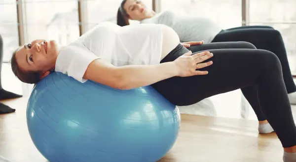 Kobiety w ciąży ćwiczą pilates na dużej, niebieskiej piłce gimnastycznej, dbając o formę i dobre samopoczucie.