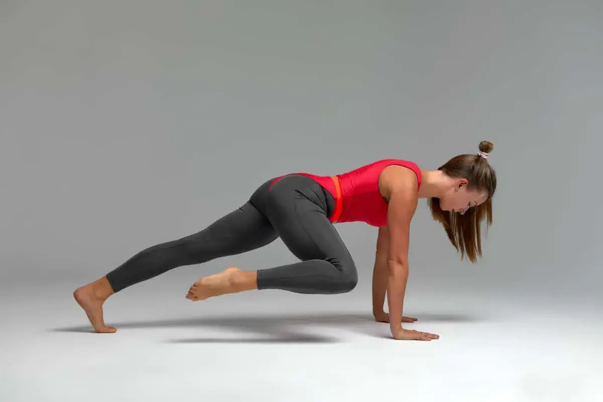 Kobieta w czerwonej bluzce i szarych legginsach wykonuje dynamiczny stretching, przyciągając jedno kolano do klatki piersiowej.