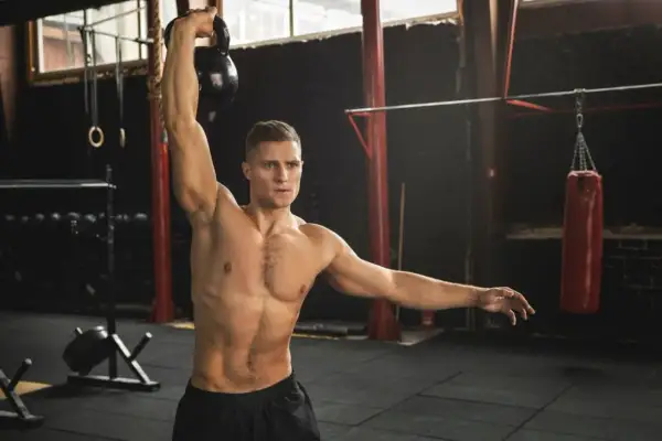 Mężczyzna wykonuje ćwiczenia na barki z kettlebell. W tle widać worek bokserski i sprzęt do treningu.