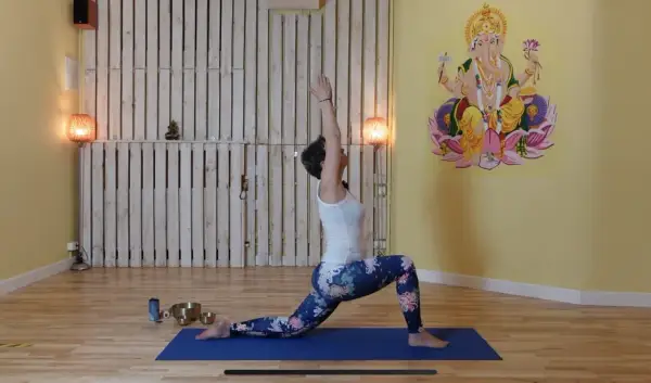 Kobieta w pozycji jogi na macie, z rękami uniesionymi ku górze. W tle obraz Ganesha i dekoracje. Czakra fioletowa symbolizuje duchowość i intuicję.